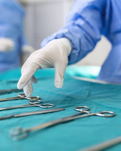 Nurse hand taking surgical instrument for group of surgeons at background operating patient in surgical theatre. Steel medical instruments ready to be used. Surgery and emergency concept